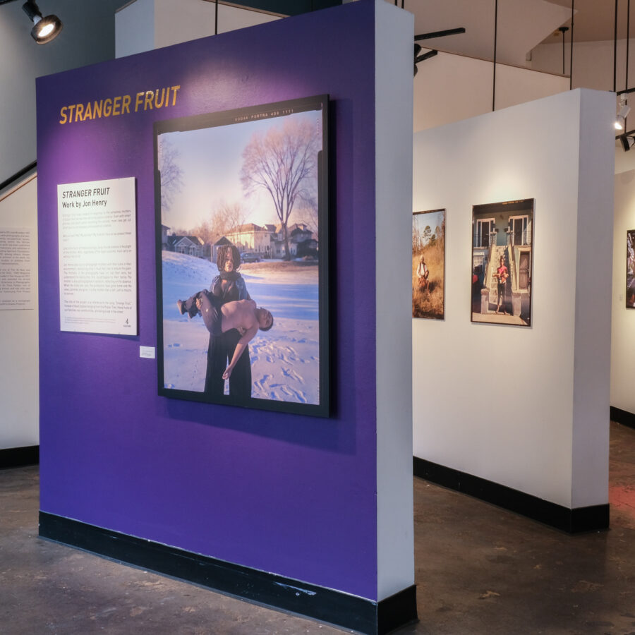 Installation photo of Stranger Fruit, work by Jon Henry. The main gallery wall of PCNW's gallery with a large photo of a black woman holding her child. The mother in the photograph has not lost her son, but understands the reality that this could happen to her family. Next to the photo is a panel with the title and artist statement for this show.